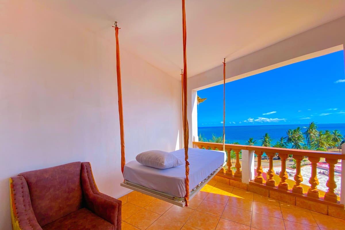 A room with a bed on a balcony, a chair, and a view of the ocean.