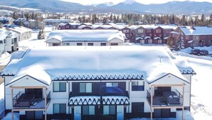 A modern multi-story building with a covered outdoor area, surrounded by snow-covered houses and mountains in the background. - 2 Mi to Fraser Tubing Hill: Condo w/ Fireplace! (Fraser)