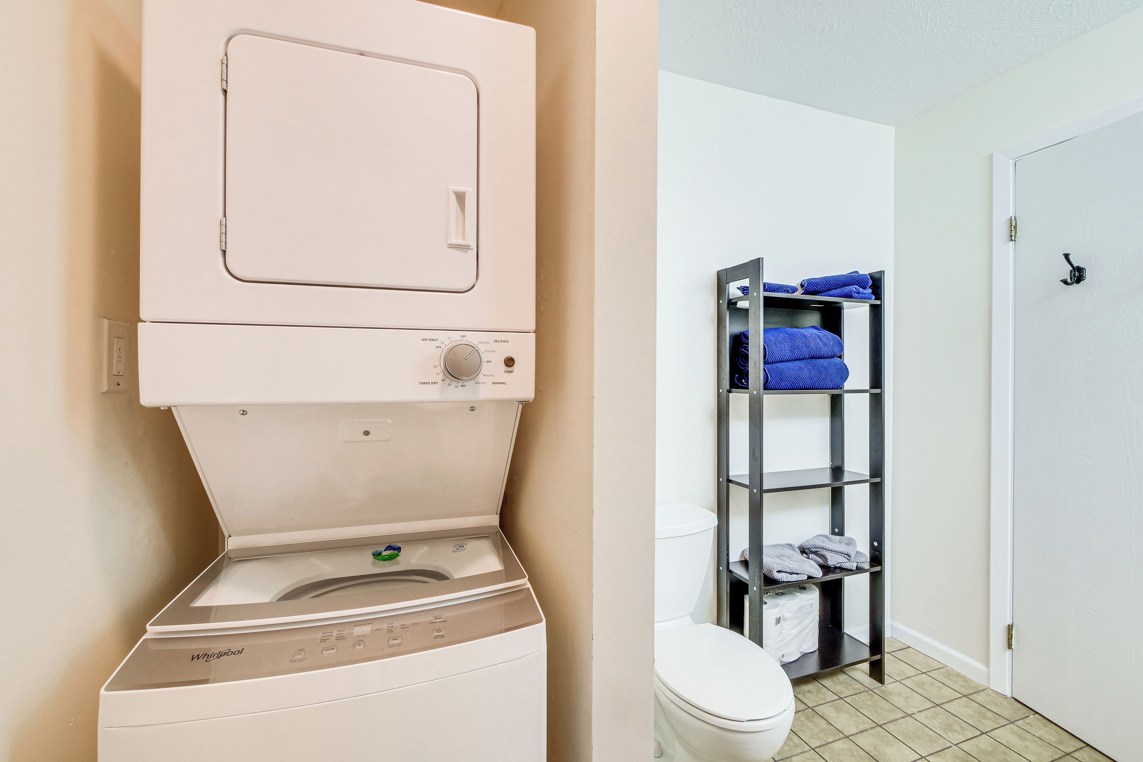 A compact laundry area with a front-loading washer and dryer, a toilet, and a metal shelf with folded towels.