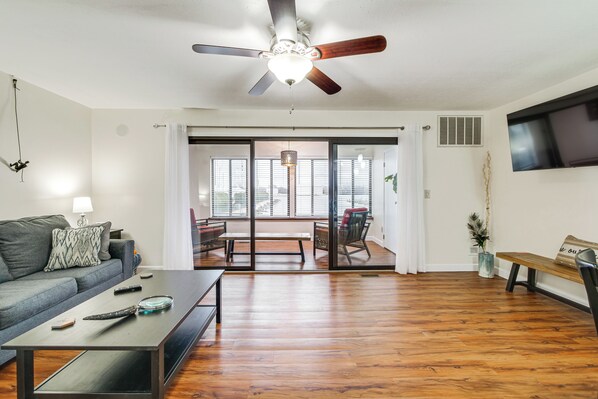 A spacious living room with a ceiling fan, a sofa, a coffee table, and a sliding glass door leading to an outdoor patio. - Lakefront Port Clinton Condo w/ Boat Dock & Slip! (Port Clinton)