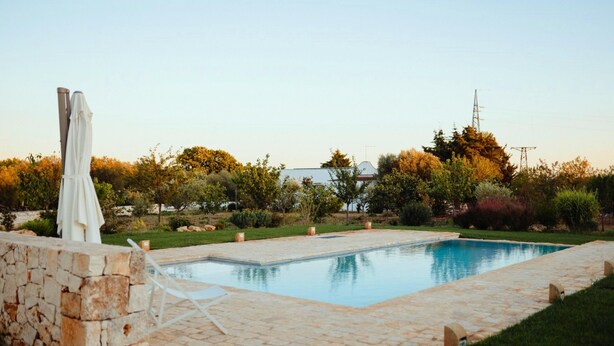 Una piscina rectangular con borde de piedra, rodeada por un muro de piedra y una zona ajardinada con árboles y arbustos.