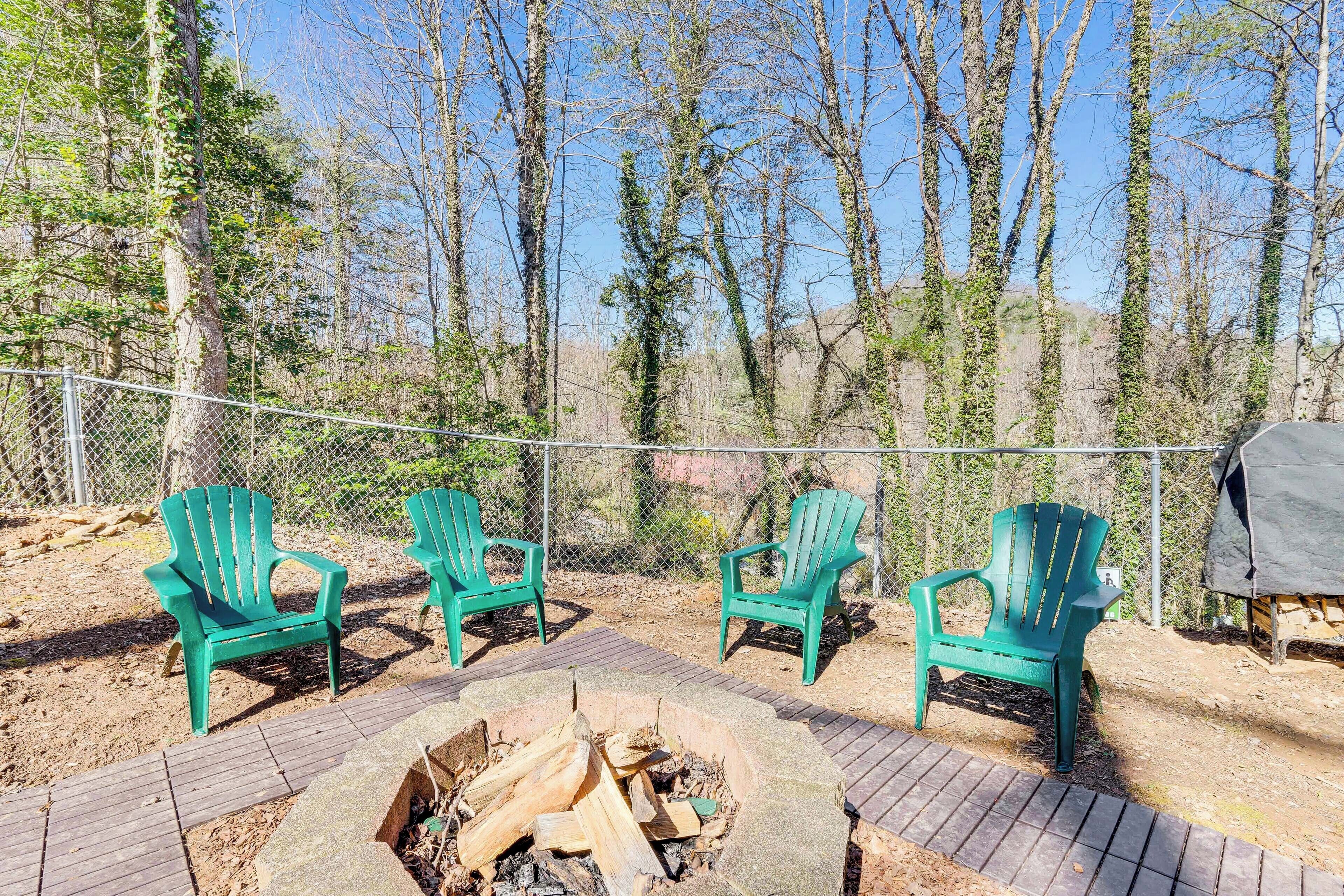 En terrasse med tre grønne Adirondack-stole, et bålsted og et grillområde, omgivet af et pigtrådshegn og træer.