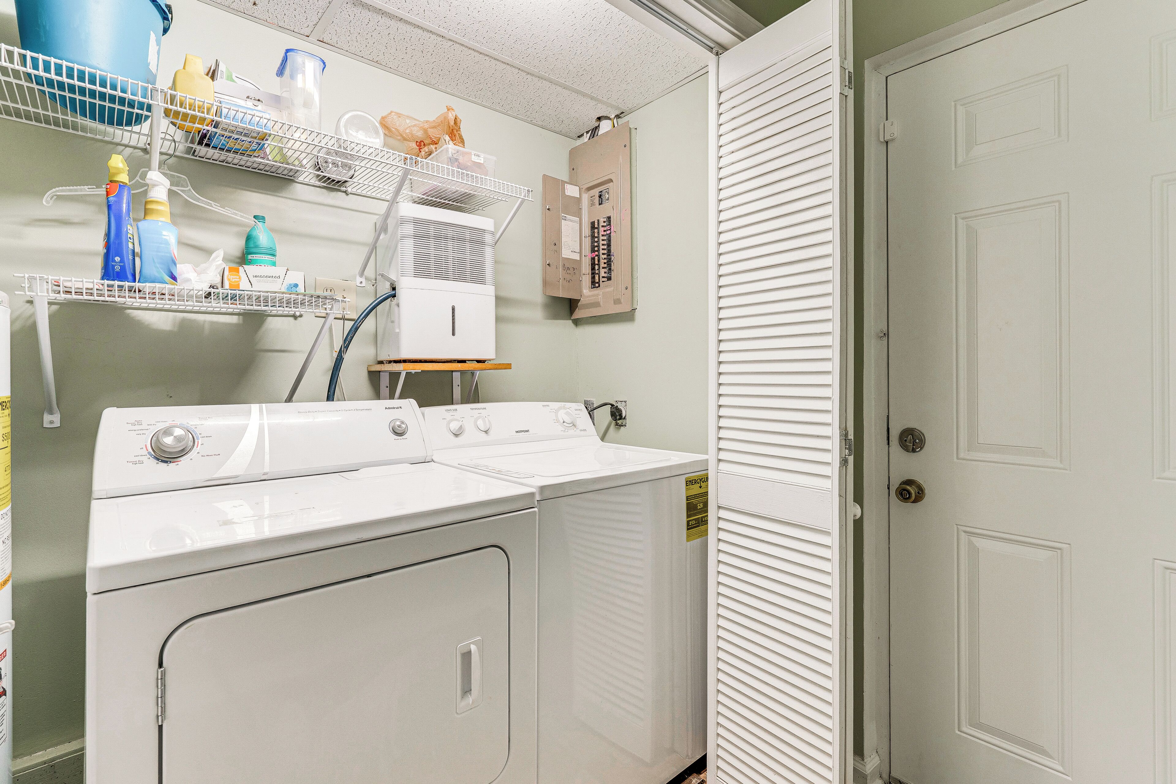 Un cuarto de lavado con lavadora y secadora apiladas, estantes con productos de limpieza y una puerta con cerradura.