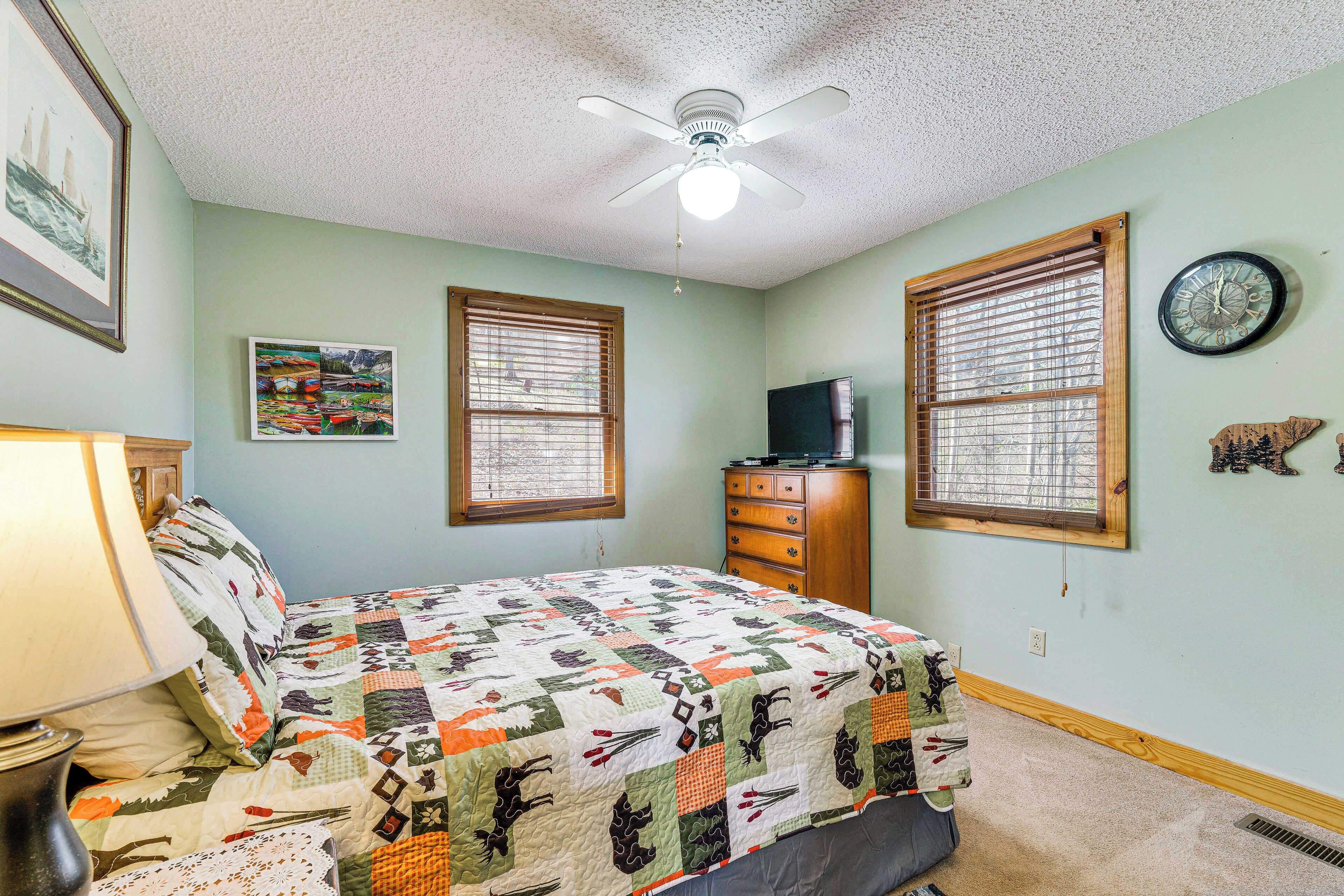 A bedroom with a bed, a ceiling fan, a dresser, a clock, and a window with blinds.