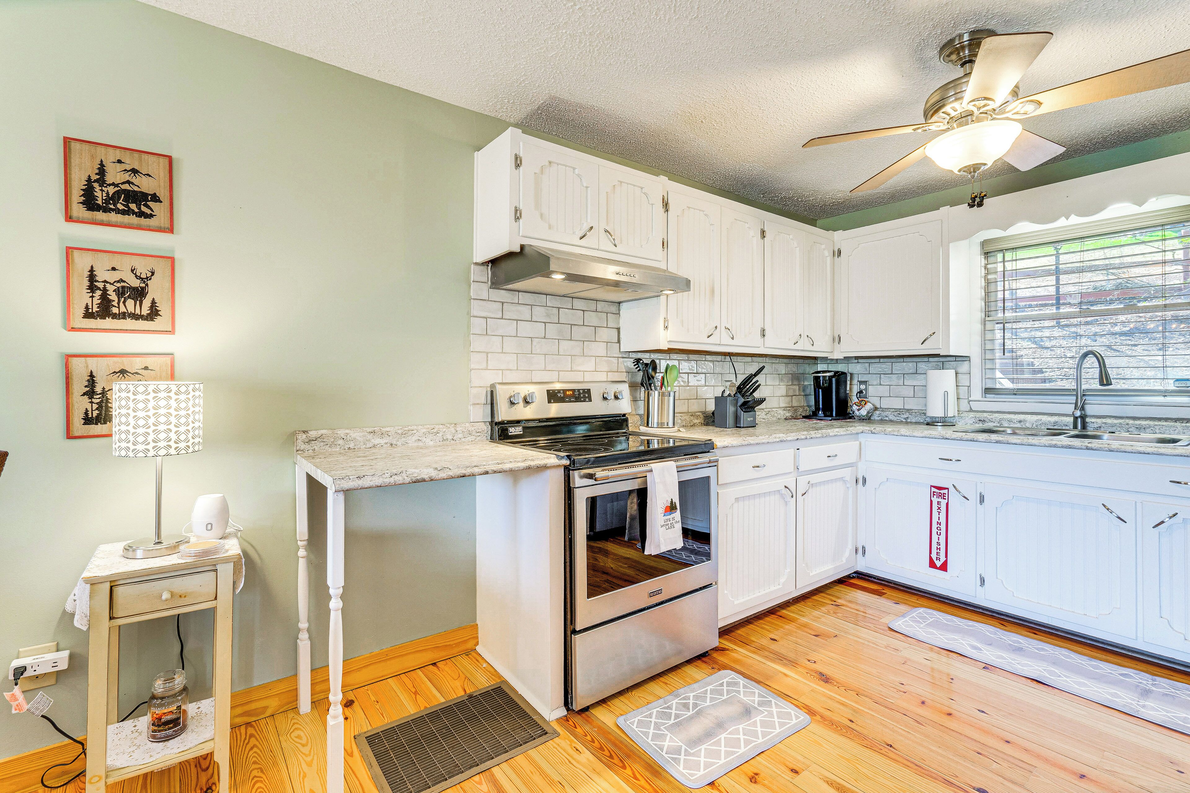 A kitchen with white cabinets, a stove, and a ceiling fan. There are framed pictures on the wall and a window with blinds.