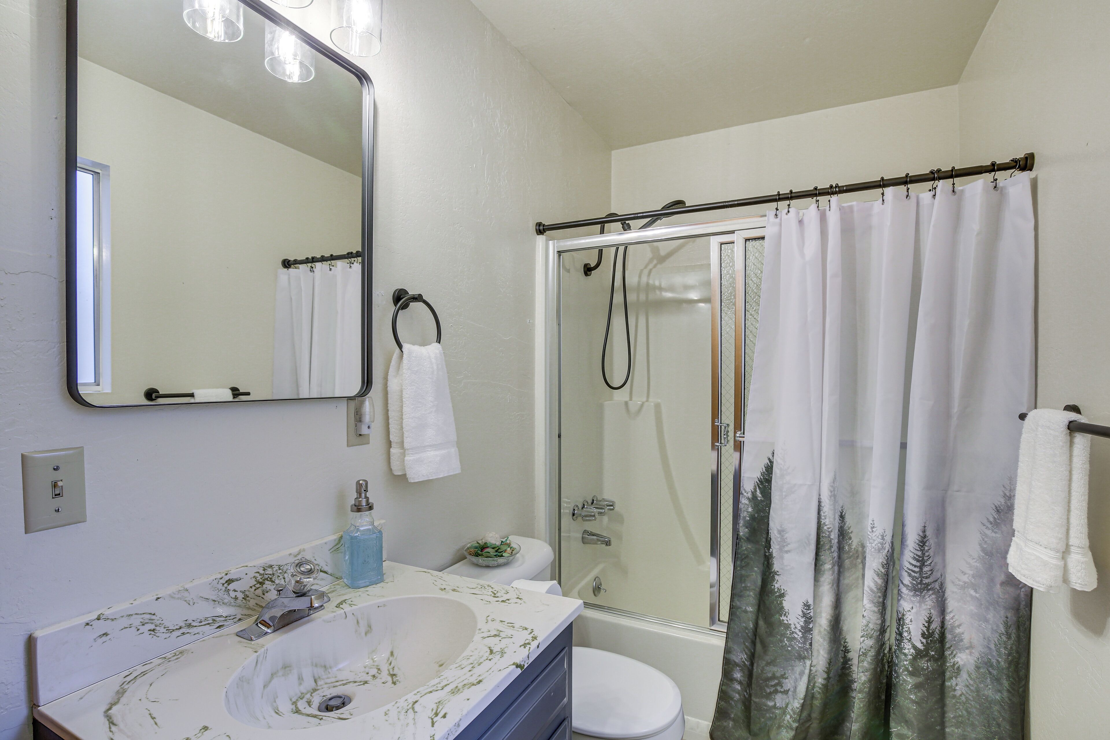 Une salle de bain avec un lavabo blanc, un miroir, une douche avec une porte en verre et un rideau orné d’un motif de forêt.