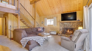 A cozy cabin interior with a fireplace, a sofa, a recliner, and a staircase leading to an upper level.