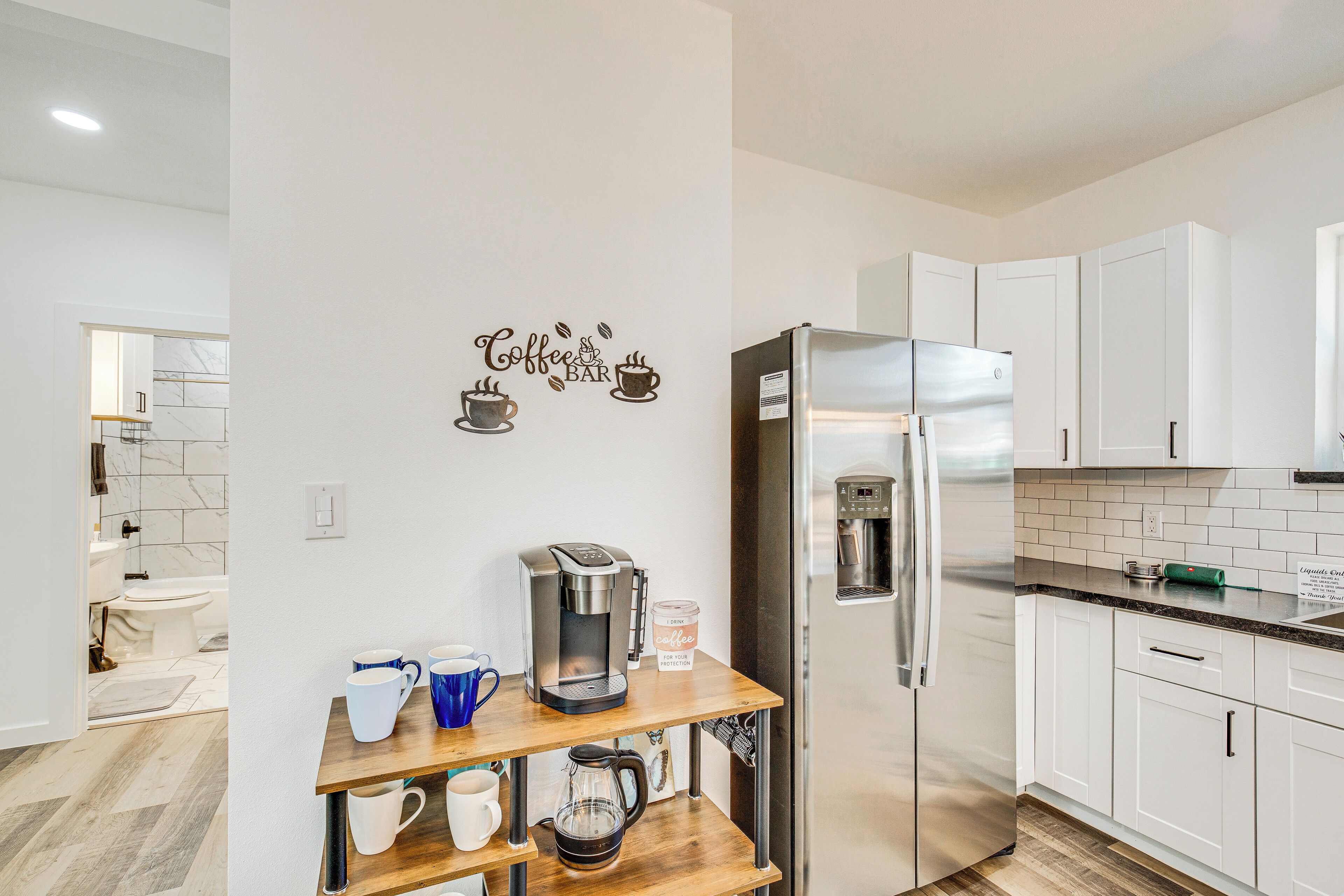 A modern kitchen with white cabinets, a stainless steel refrigerator, a coffee maker, and a coffee grinder on a wooden shelf.