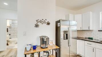 A modern kitchen with white cabinets, a stainless steel refrigerator, a coffee maker, and a coffee grinder on a wooden shelf.