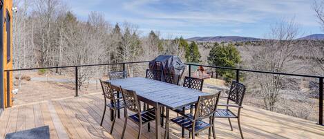 A wooden deck with a table and chairs, a barbecue grill, and a view of trees and hills.