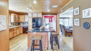 A spacious kitchen with wooden cabinets, a central island with stools, and a view into the dining area. - Riverside Texas Retreat w/ Wraparound Porch! (Thorndale)