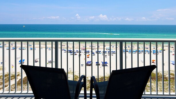 A balcony with two chairs overlooking a beach with umbrellas and people.
