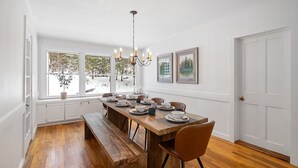 Un comedor con una mesa de madera dispuesta para cuatro personas, paredes blancas y vistas a un paisaje nevado a través de amplios ventanales.