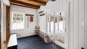 Una habitación con techo de madera, un banco, un perchero con una mochila, una puerta que da al exterior y una ventana con vistas a un paisaje nevado.