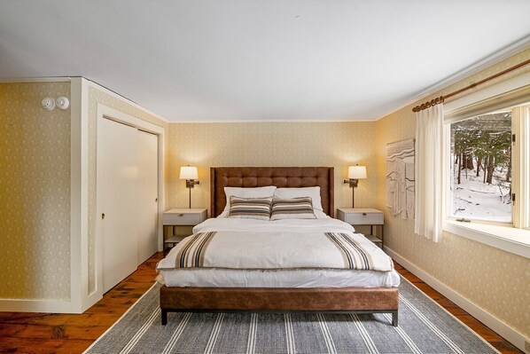Ein Schlafzimmer mit einem großen Bett, zwei Nachttischen, einem Fenster mit Blick auf eine verschneite Landschaft und einem gemusterten Teppich.