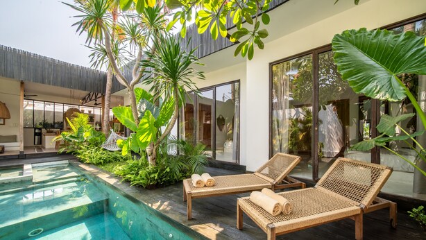 A modern outdoor pool area with two wicker lounge chairs, surrounded by lush tropical plants and large windows. - Luxueuse Villa Yogisurf 2 Canggu Piscine & Jacuzzi (Kerobokan)