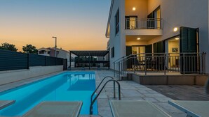 A swimming pool with lounge chairs, a covered seating area, and a building with balconies and windows. - Villa Kristina (Nin)