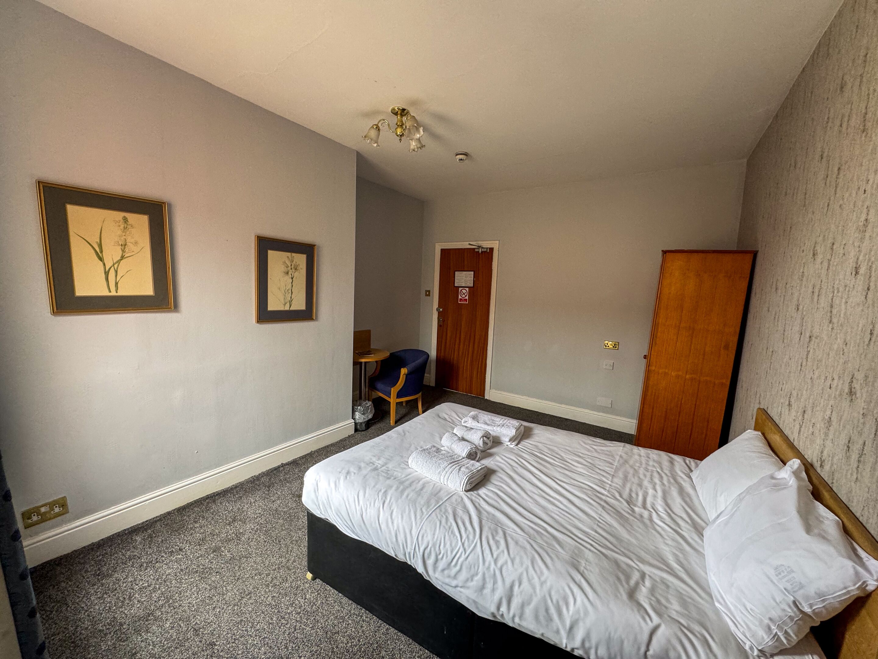 A hotel room with a bed, two framed pictures on the wall, a chair, a desk, and a wardrobe.