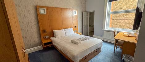 Un dormitorio con cabecera de madera, cama con ropa blanca, una mesita de noche, un escritorio con silla y una ventana con vista a un muro de ladrillo.