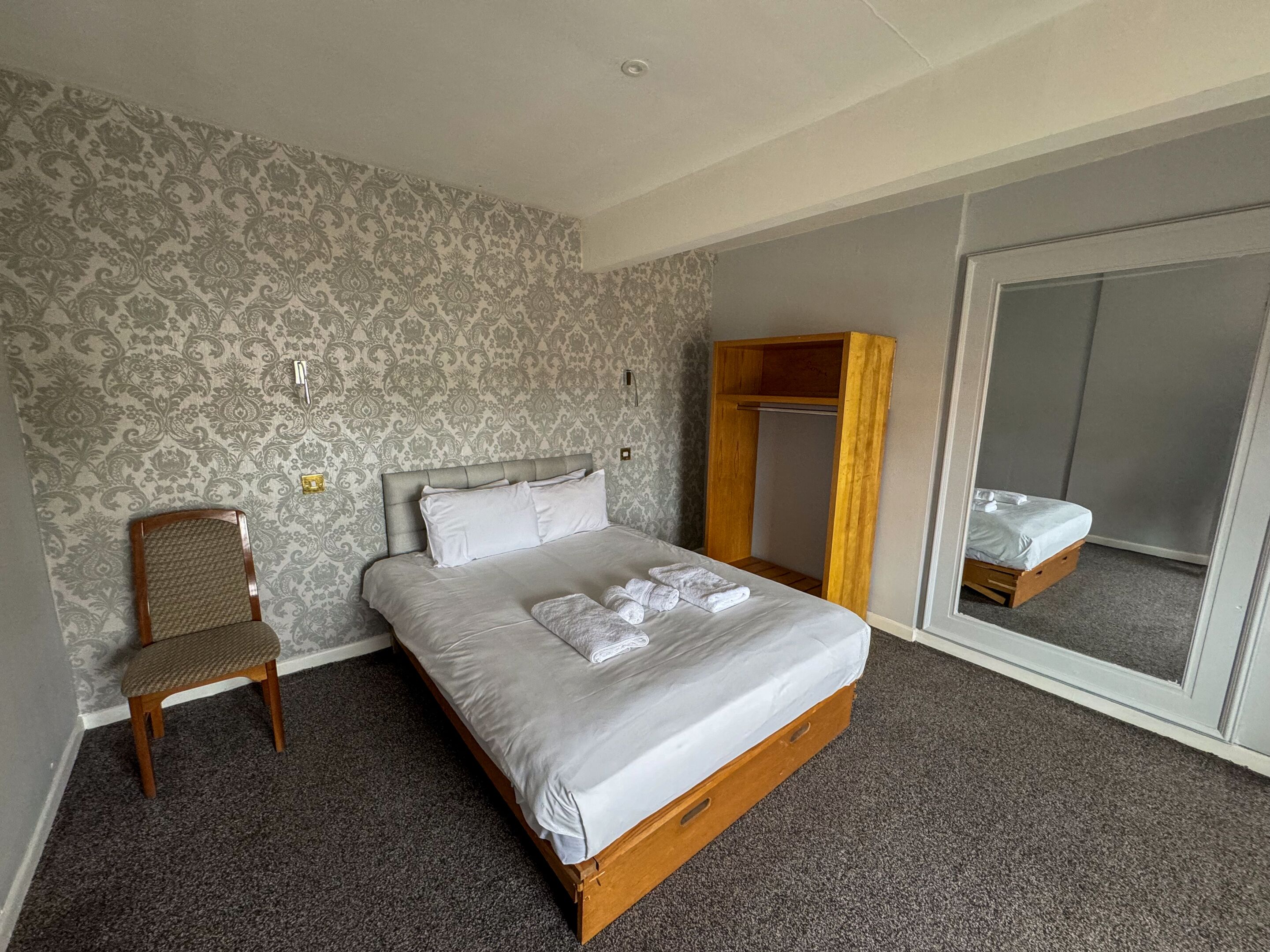 Una camera d'albergo con un letto, una sedia in legno, un armadio e uno specchio grande.