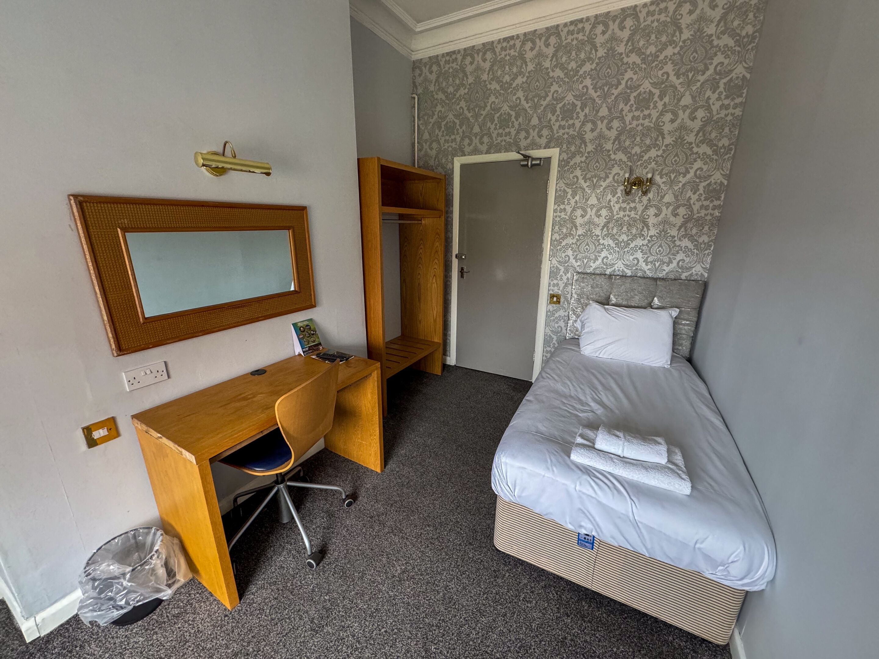 A hotel room with a bed, a desk, a chair, a mirror, and a wardrobe.