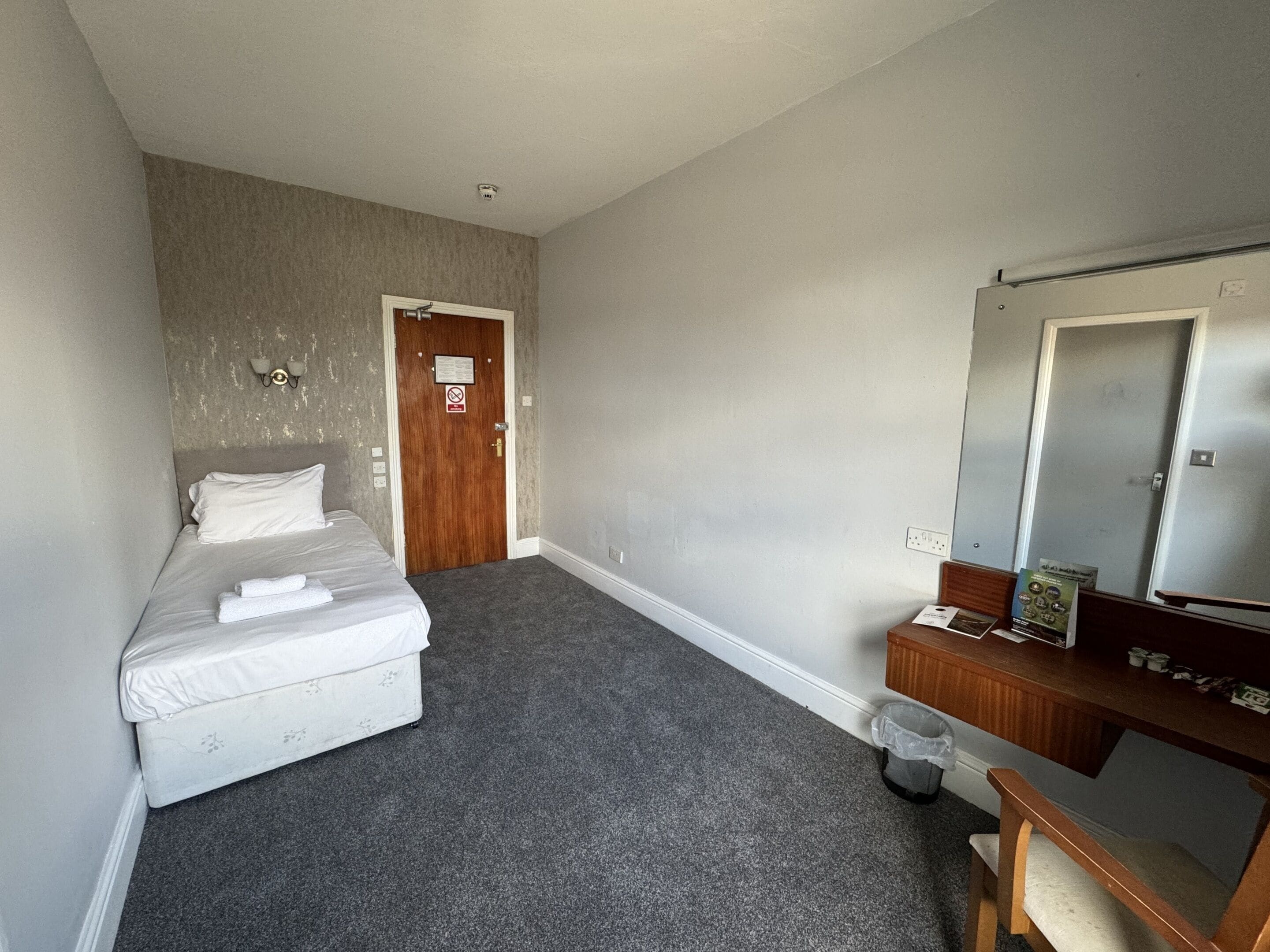 A hotel room with a bed, a wooden dresser, a mirror, and a door with a no-smoking sign.