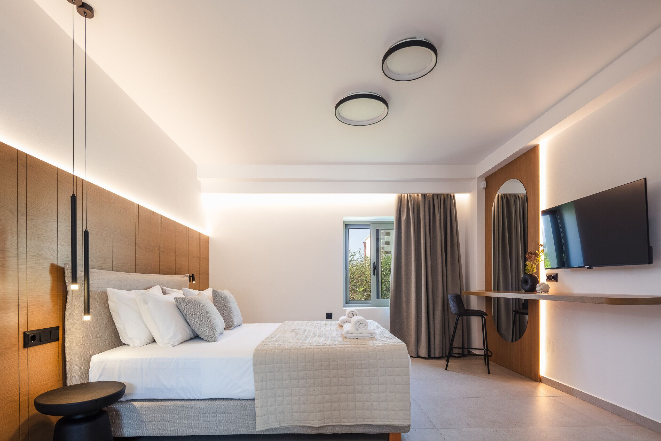 Ein modernes Hotelzimmer mit einem großen Bett, einem an der Wand montierten Fernseher, einem Schreibtisch mit Stuhl und einem Fenster mit Vorhängen.