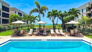 Un espace piscine agrémenté de chaises longues et de parasols, entouré de palmiers et offrant une vue sur l’océan.