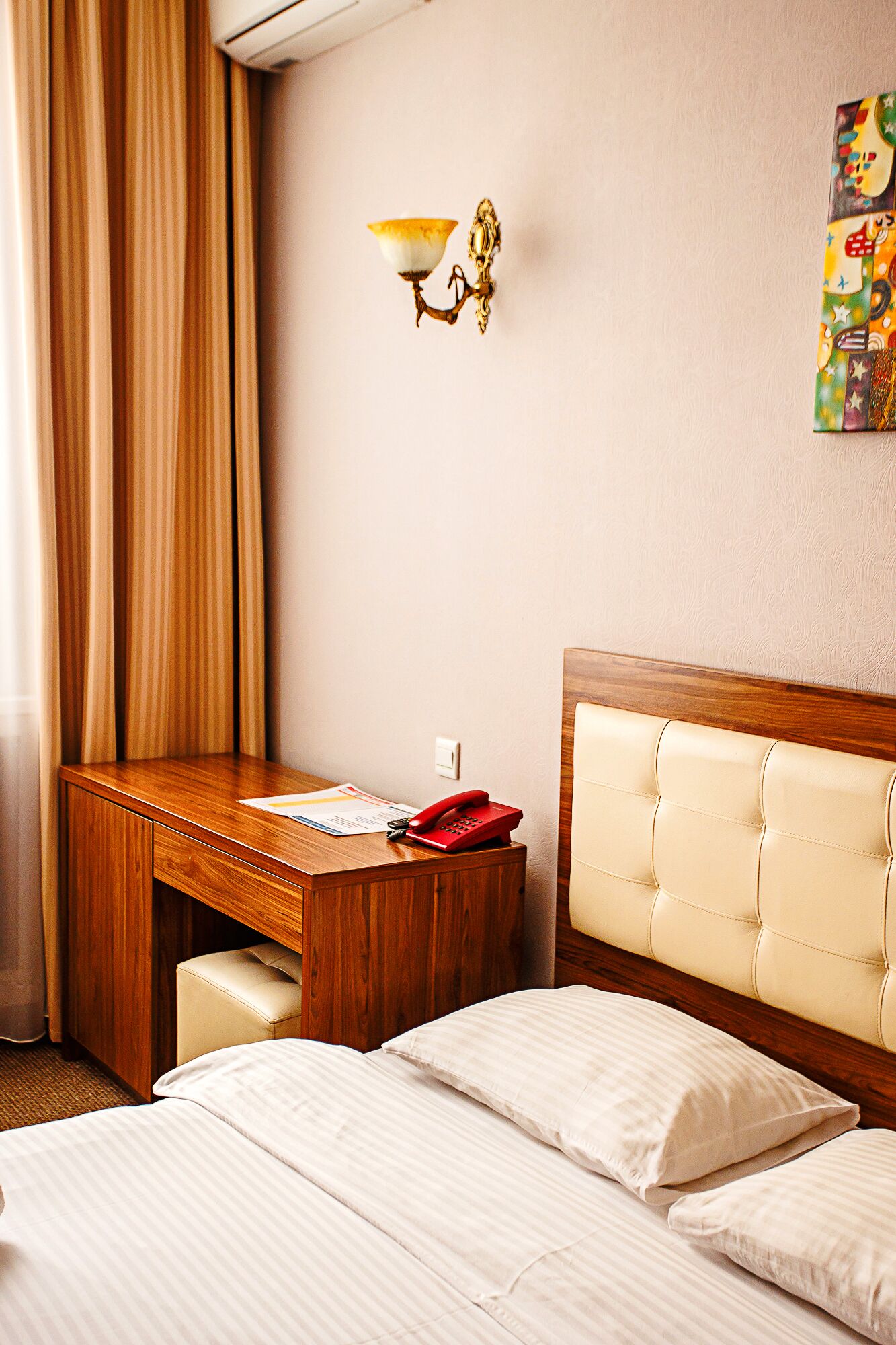 Habitación de hotel con un escritorio de madera, un teléfono rojo, cabecero acolchado y una lámpara de pared.
