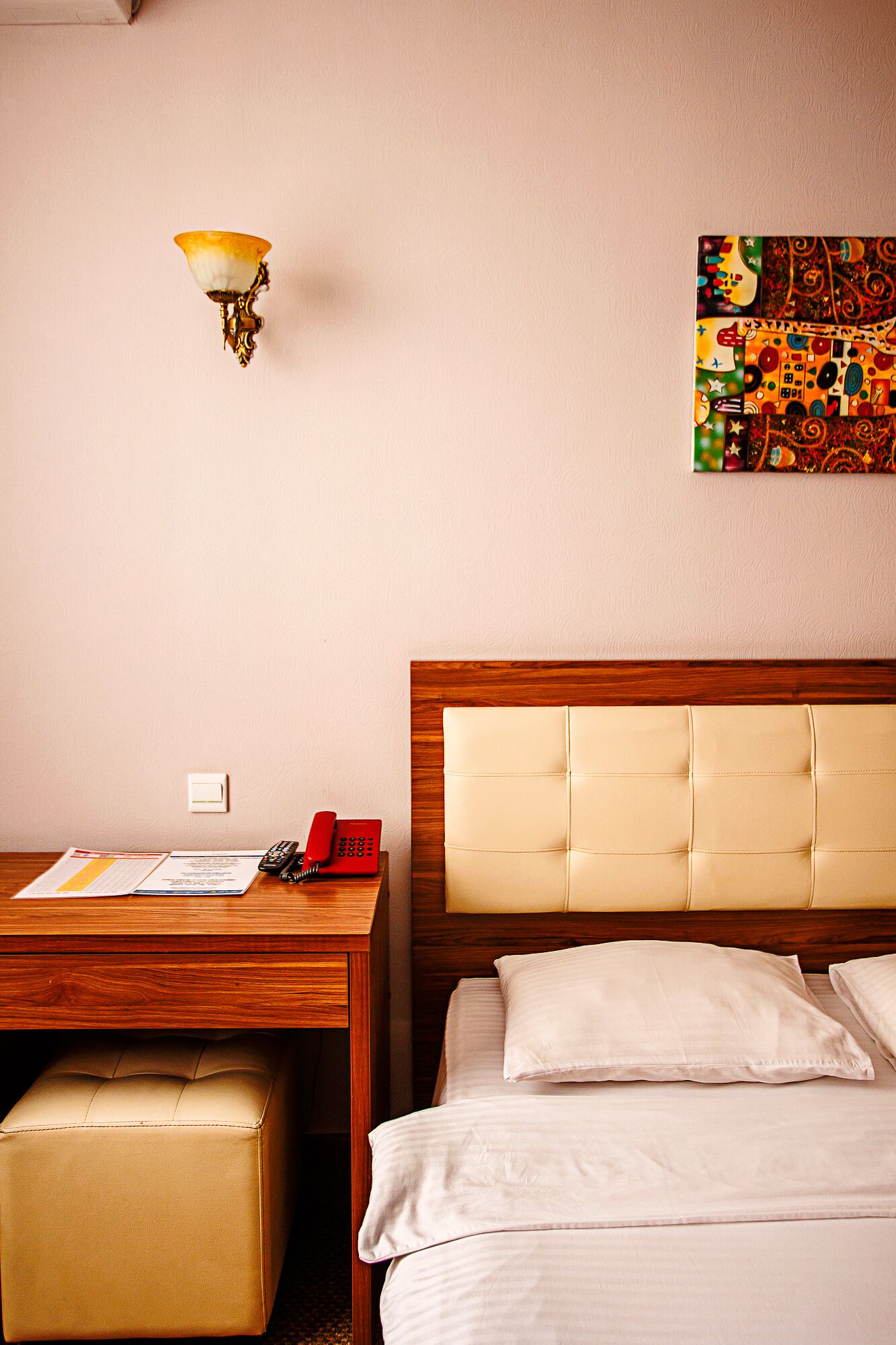 Habitación de hotel con un escritorio de madera, teléfono, lámpara, cama con cabecera y una obra de arte colorida en la pared.