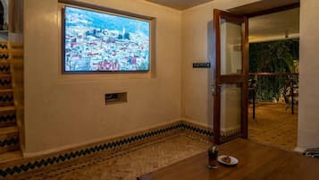 Una habitación con un ventanal que muestra un paisaje urbano, una mesa de madera con un vaso de agua y un plato de comida, y un suelo con diseño.