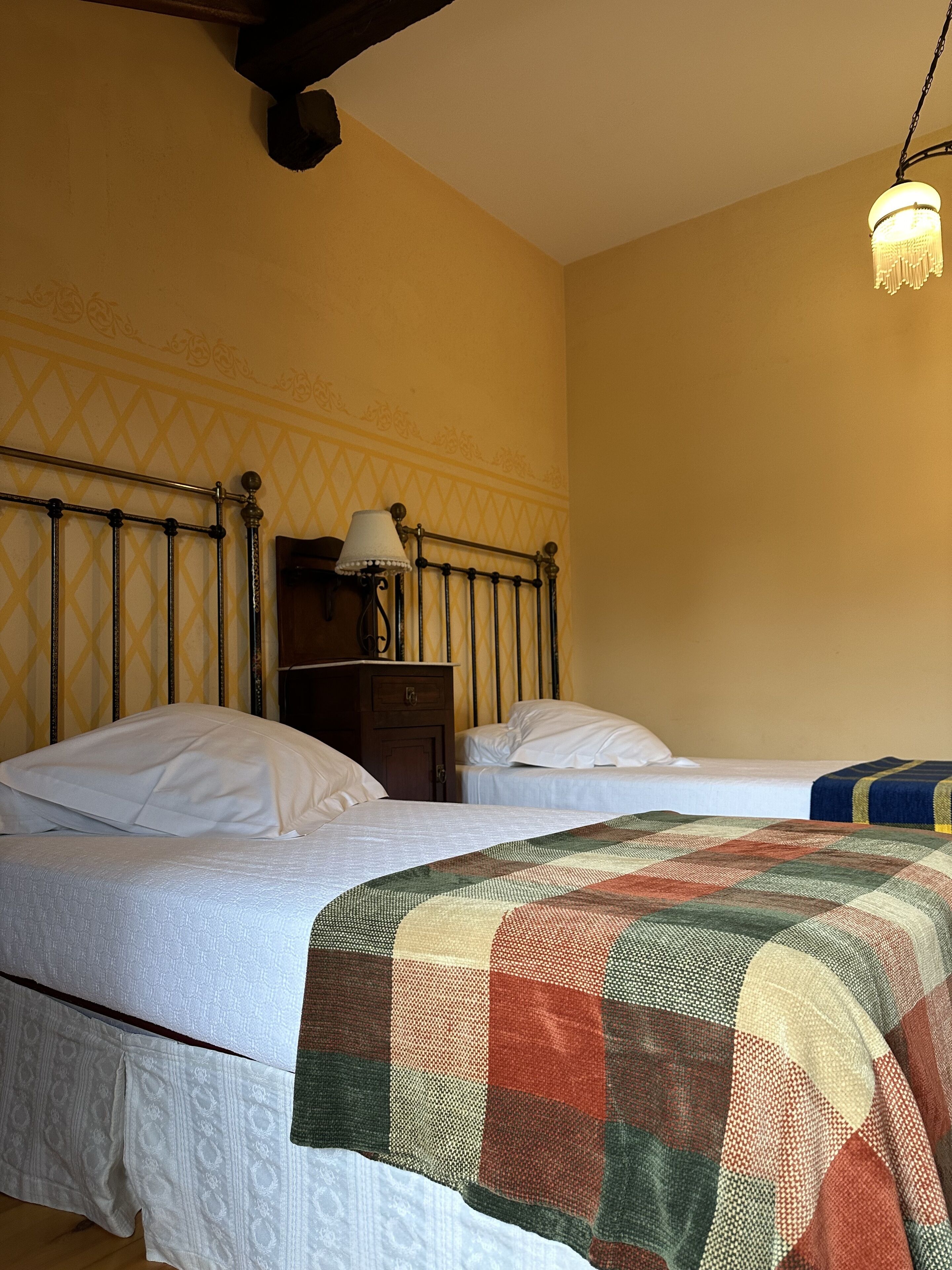 A bedroom with two beds, a wooden headboard, a nightstand with a lamp, and a patterned wall.
