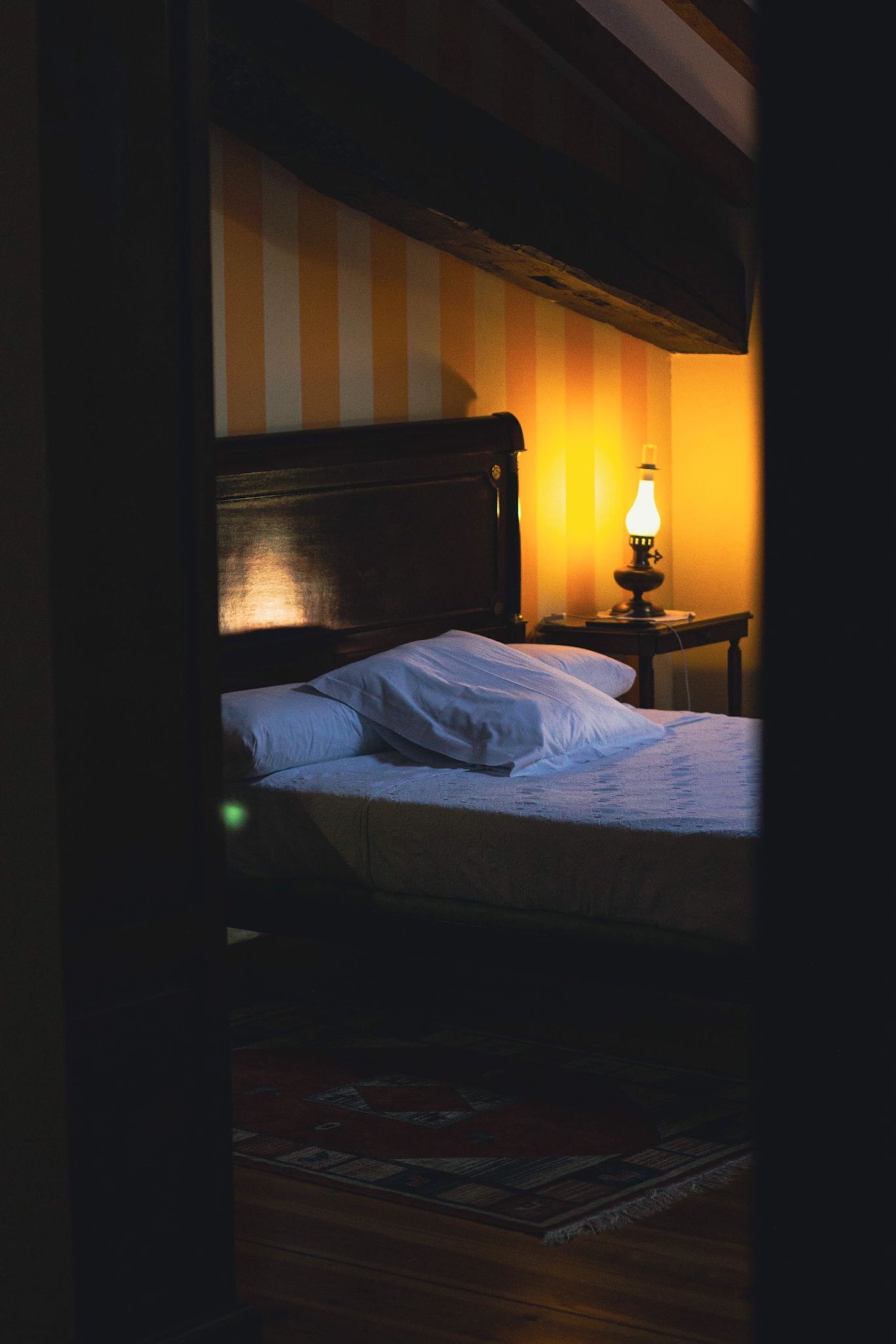 A dimly lit room with a bed, a nightstand with a lamp, and a striped wall.