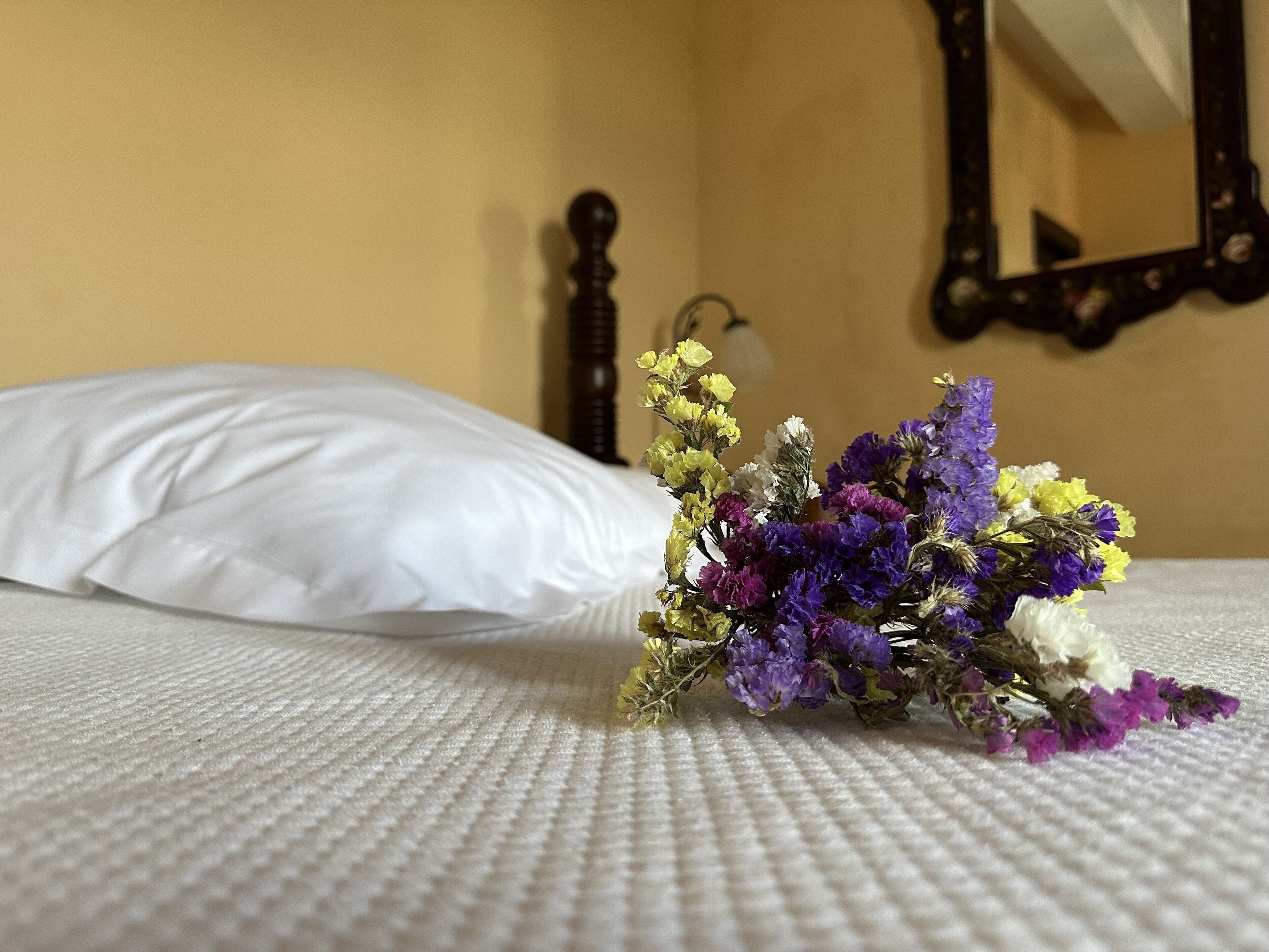 Una cama con un almohadón blanco y un ramo de flores sobre una colcha blanca con textura.