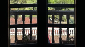 Une vue à travers une fenêtre ornée d’un balcon, donnant sur un paysage verdoyant avec des arbres et des collines.