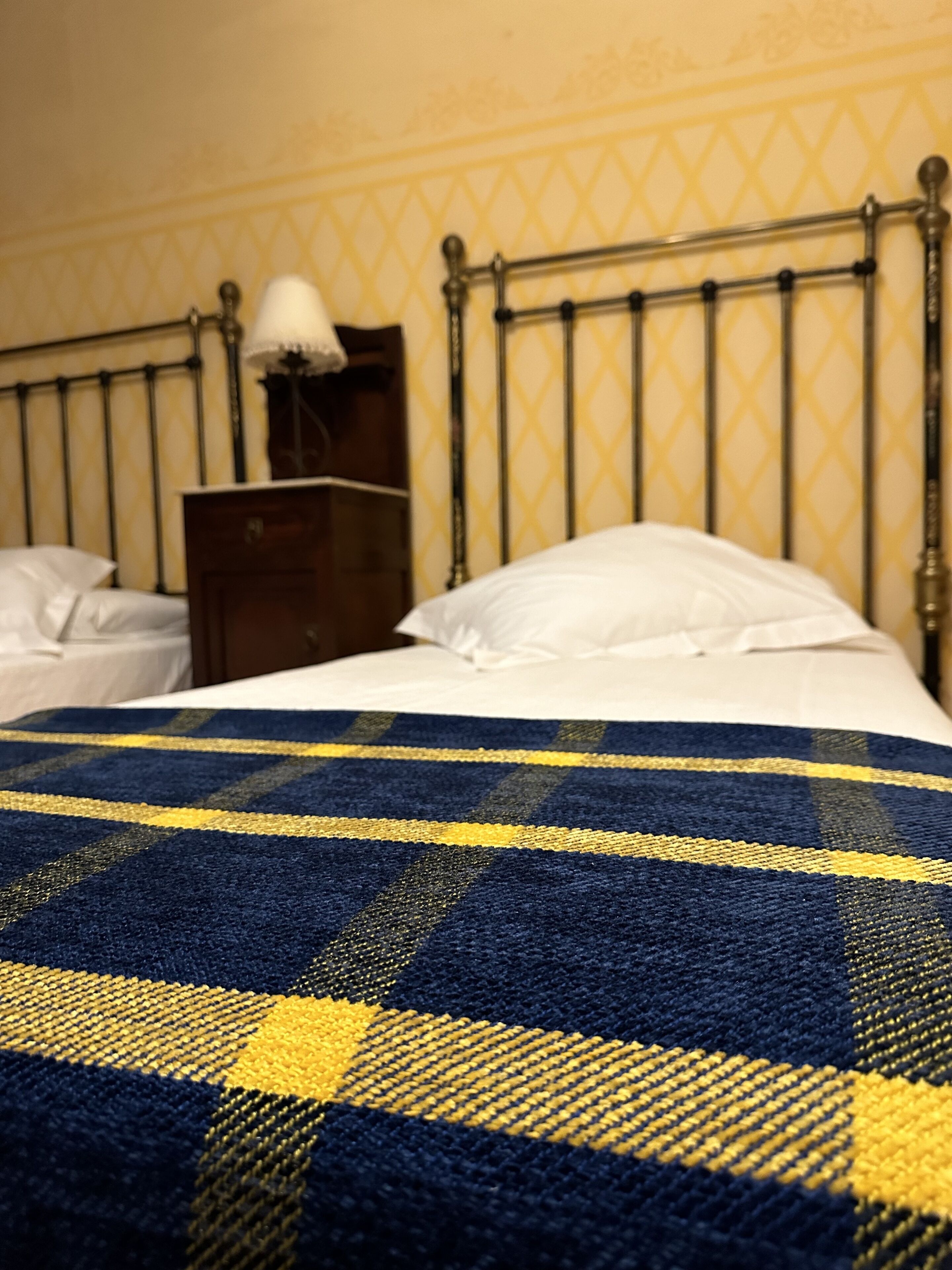 Un lit recouvert d’une couverture à carreaux bleus et jaunes, une table de chevet en bois avec une lampe et une tête de lit en métal.