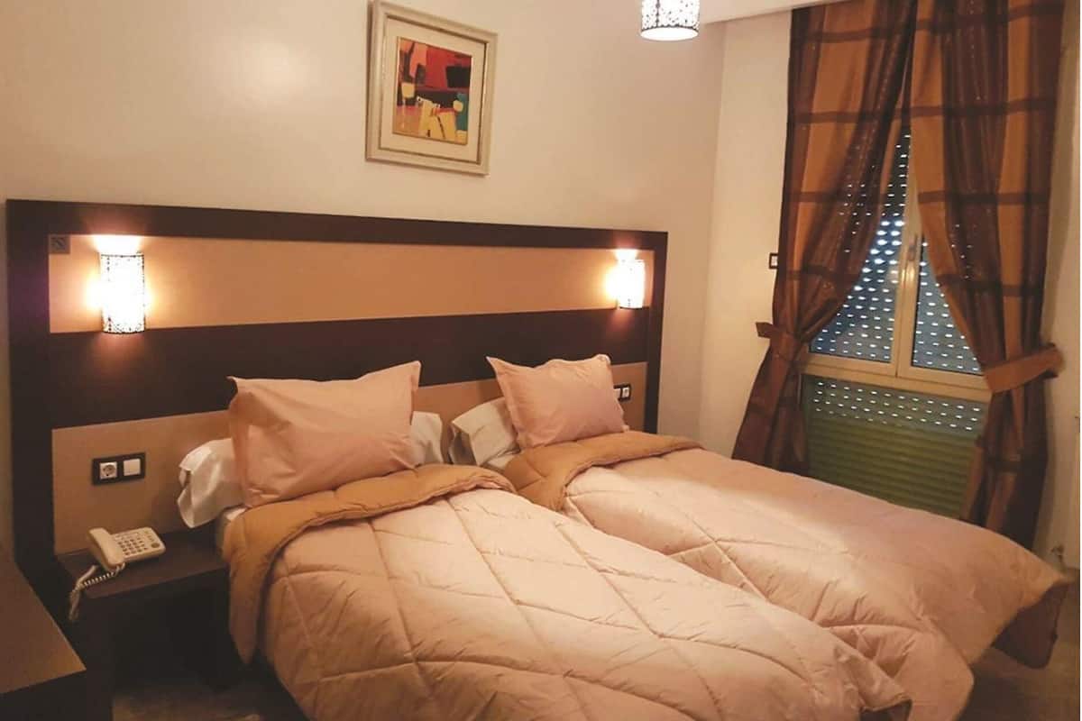 Una cama doble con edredón acolchado, lámparas de noche, teléfono y ventana con cortinas.