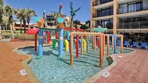 Un espace de jeux aquatiques avec des structures colorées et une piscine, entouré d’un chemin pavé et de palmiers.