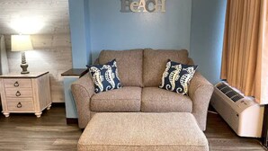 Un salon avec un canapé beige, deux coussins assortis, une petite table d’appoint avec une lampe et un climatiseur fixé au mur.