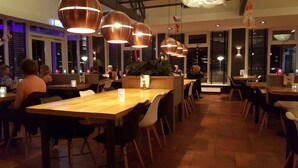 Un restaurante con mesas y sillas de madera, lámparas colgantes de cobre y una planta central.