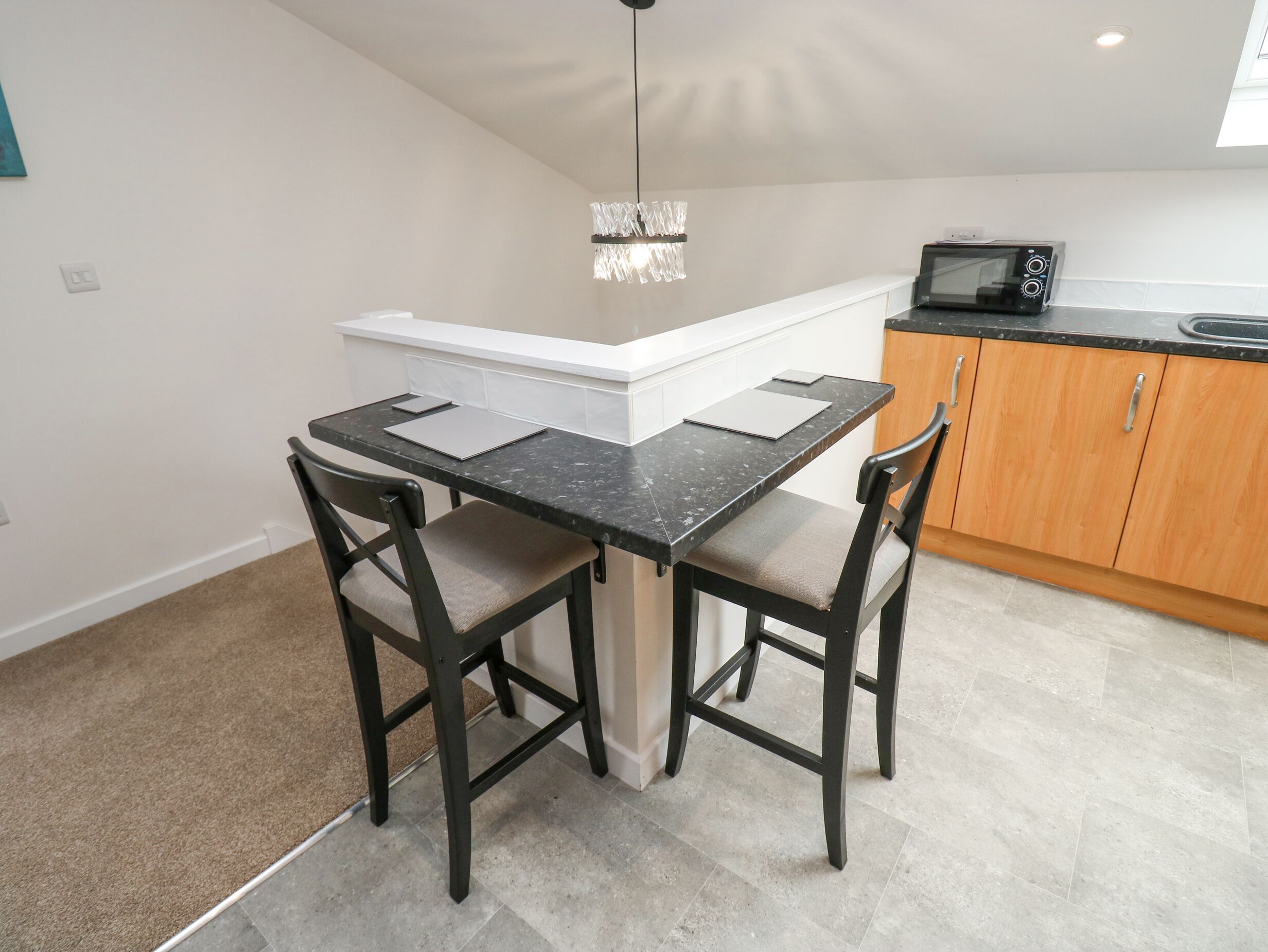 A compact kitchen with a dark marble countertop, two chairs, a small island, a microwave, and a pendant light.