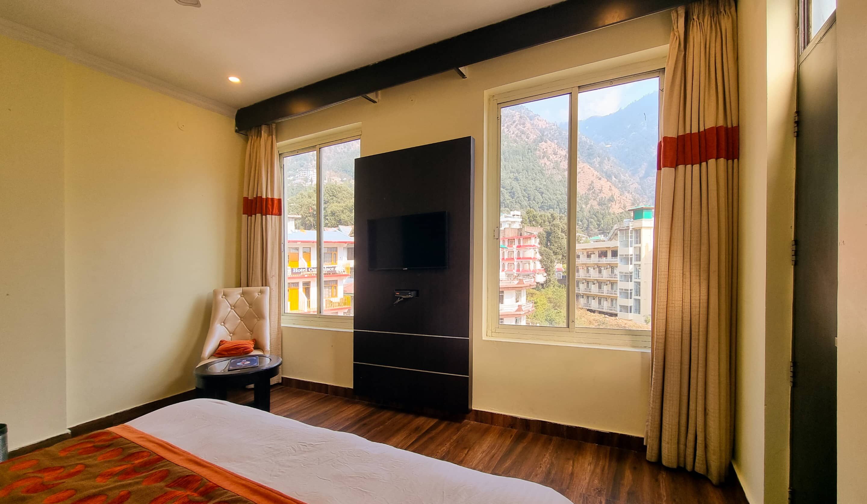 A hotel room with a bed, a TV, a small table, and a chair. There are windows with curtains and a view of buildings and mountains outside.