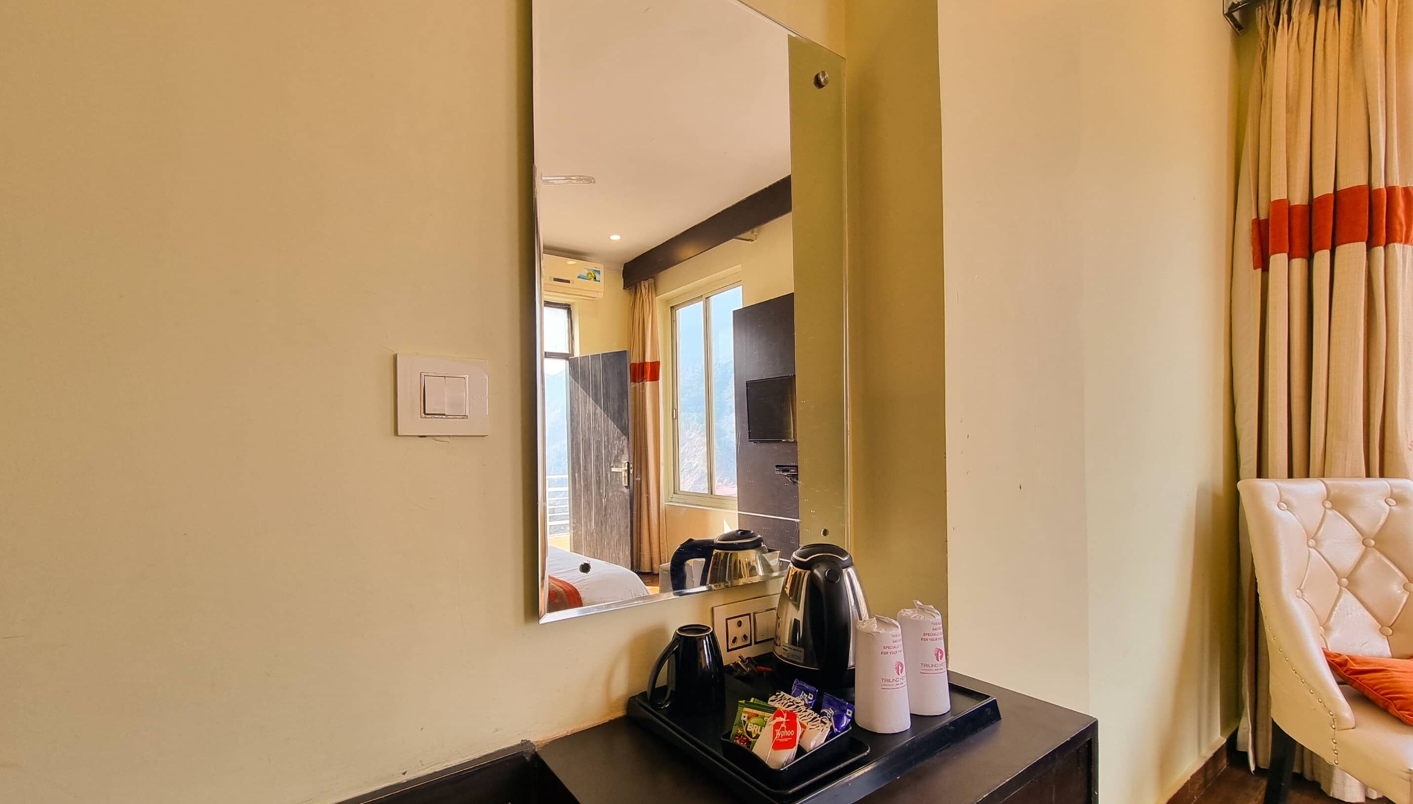 Une chambre d’hôtel dotée d’un grand miroir, d’un coin café avec une bouilloire et des tasses, et d’un fauteuil moelleux.