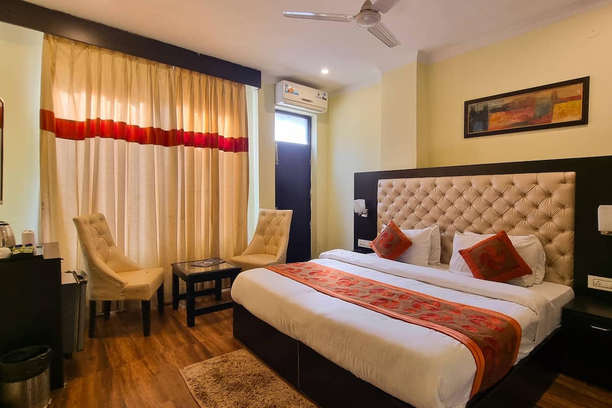 A hotel room with a bed, two chairs, a small table, an air conditioning unit, and a painting on the wall.