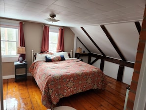 A bedroom with a sloped ceiling, a bed with floral bedding, a nightstand with a lamp, and a window with curtains. - Lizzie's Place, A Farmhouse retreat in the heart of Vermont's Northeast Kingdom. (Brunswick)