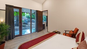 Ein modernes Schlafzimmer mit einem großen Bett, einem Holzsessel, einem kleinen Tisch und Blick auf einen Garten durch Glastüren.