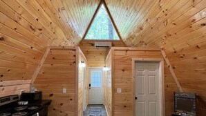 Interior rústico com teto inclinado e claraboia. Cozinha visível à esquerda, com fogão e armários. Uma porta é vista à direita, e uma lixeira está em primeiro plano.
