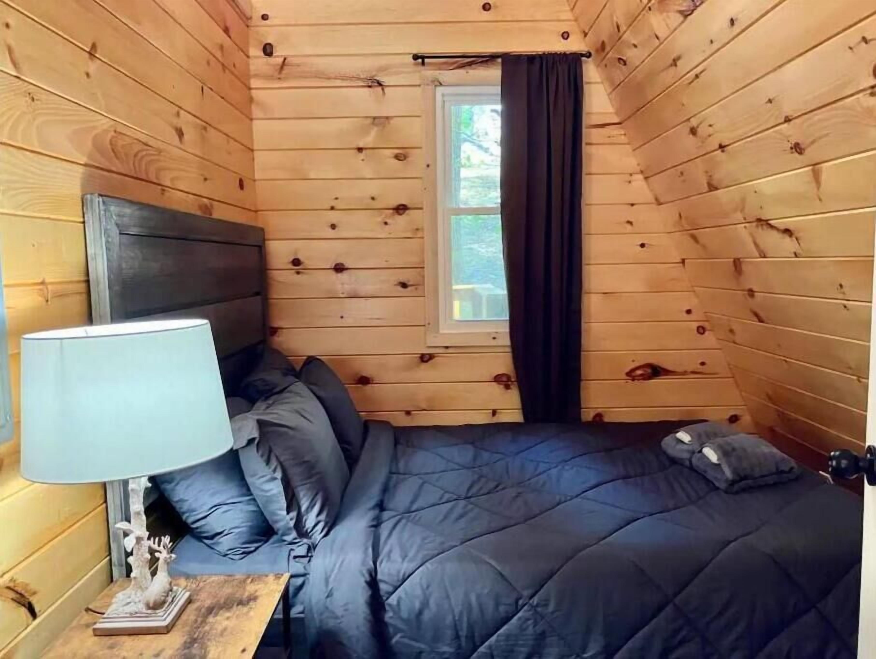 A wooden cabin room with a bed, a nightstand, a lamp, and a window with curtains.
