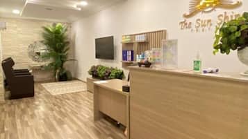 A reception area with a wooden reception desk, a seating area with a chair, a wall-mounted TV, and a wall with a sun emblem and text.