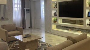 A modern living room with a large flat-screen TV, comfortable seating, and a decorative shelf. - The Palms Hotel (Abuja)